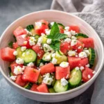 A fresh and vibrant bowl of Watermelon Salad With Cucumber And Feta, garnished with mint, featuring cubes of watermelon, cucumber, and feta.