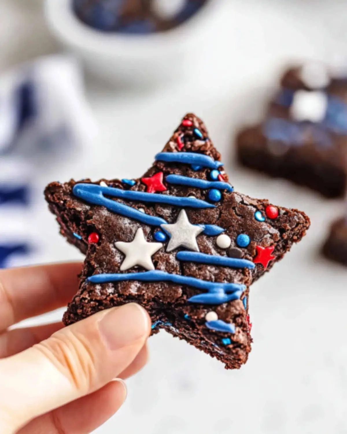 Hand holding a bite-ready star brownie with blue drizzle and silver star sprinkles, festive Fourth of July dessert in focus.