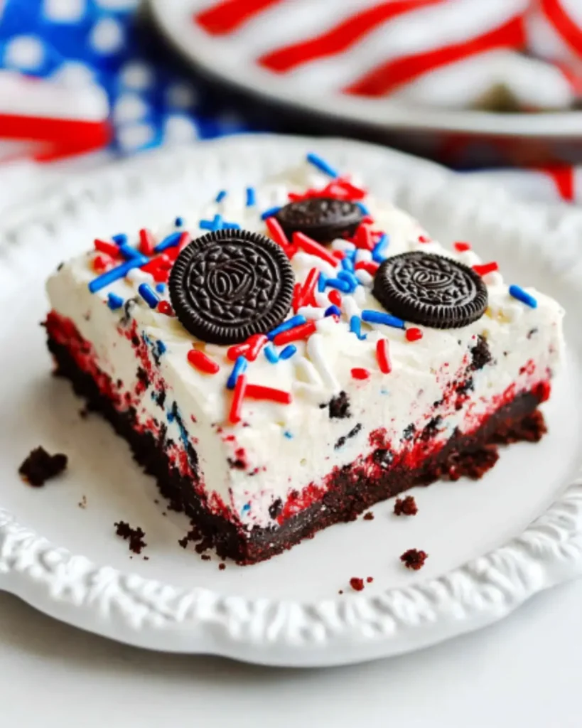 No-bake fireworks Oreo cheesecake bar topped with crushed Oreos and red, white, and blue sprinkles on a white plate, Fourth of July dessert.