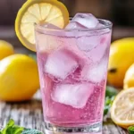Glass of pink Himalayan salt drink with ice cubes, lemon slice, and fresh mint on a rustic wooden table