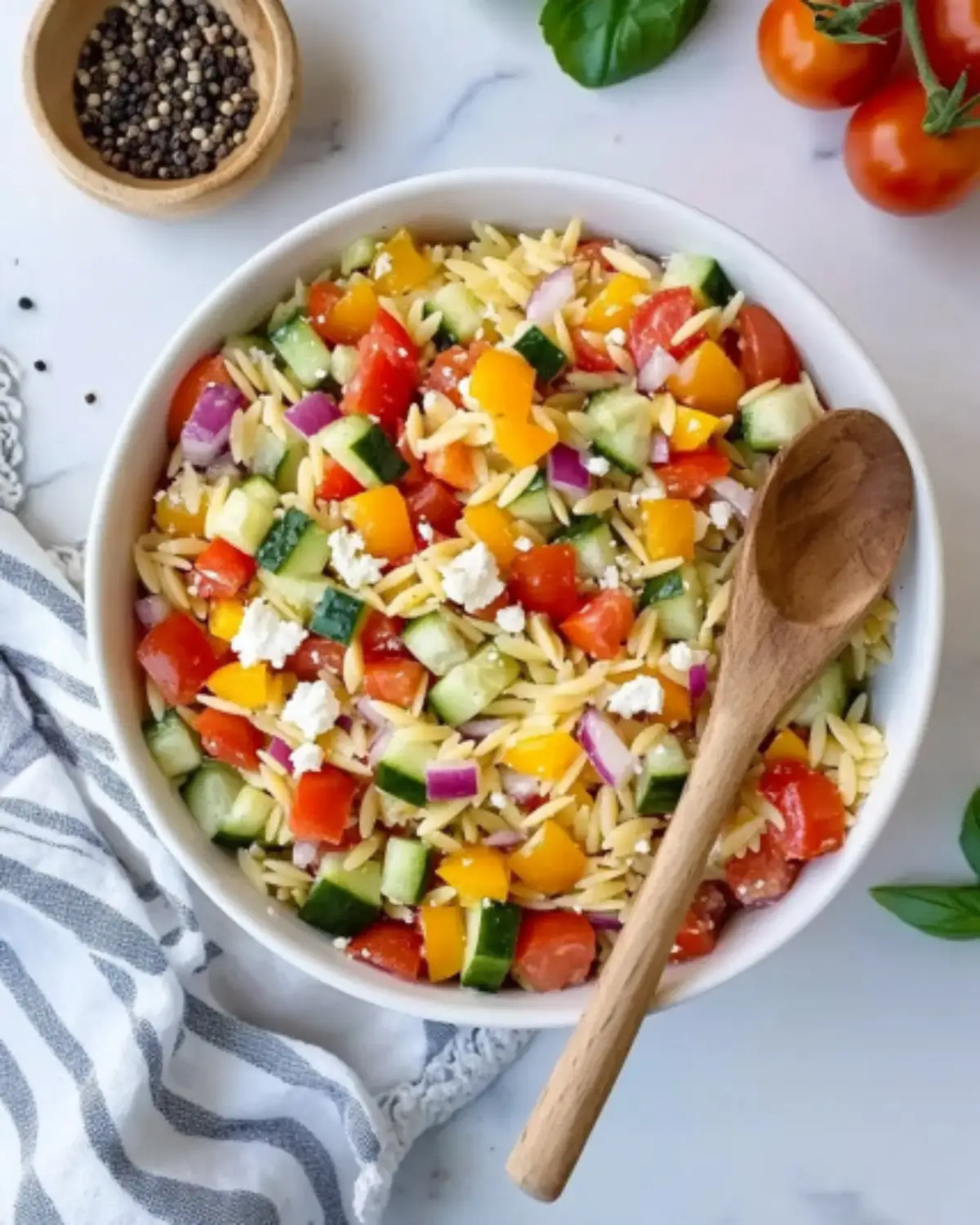Top-down view of Cold Pasta Salad with orzo, tomatoes, cucumbers, peppers, and feta in a white bowl, styled with fresh basil and striped napkin.