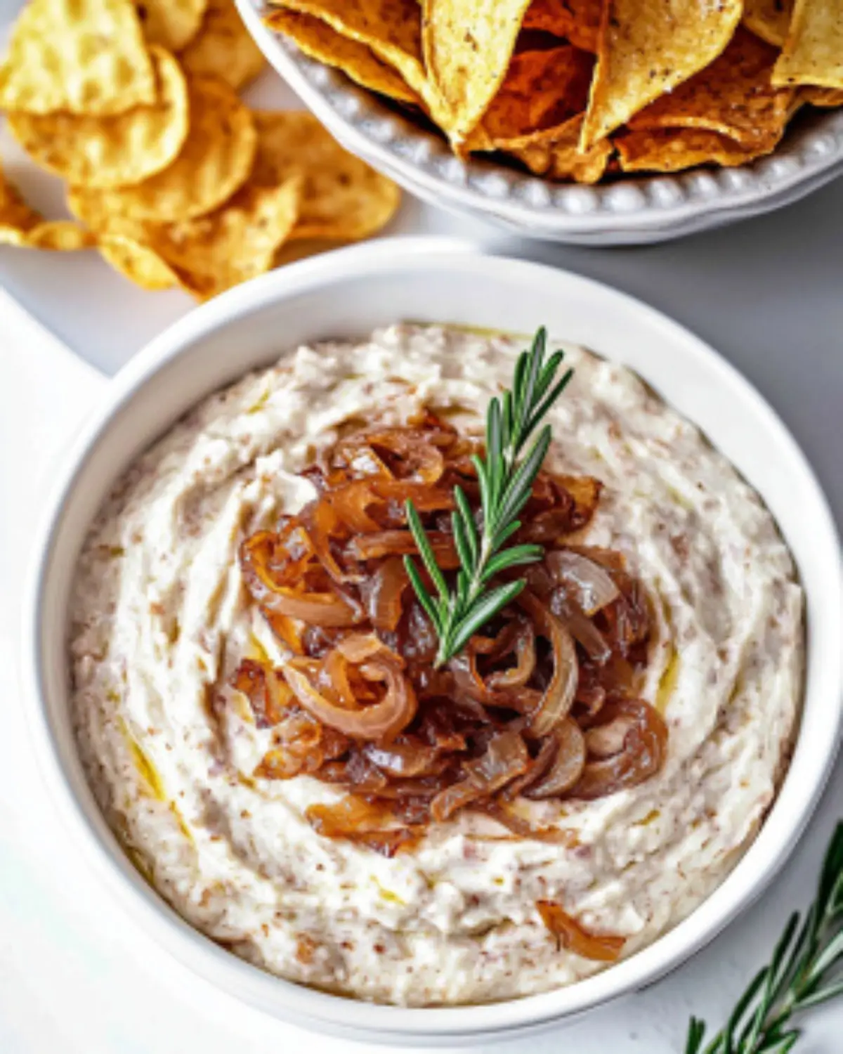 White bowl of sour cream-based Onion Dip Recipe topped with caramelized onions and rosemary, surrounded by tortilla chips.