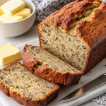 Freshly baked banana bread loaf sliced on a white marble board with butter on the side