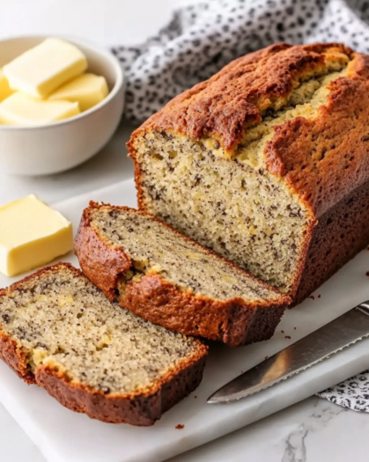 Freshly baked banana bread loaf sliced on a white marble board with butter on the side