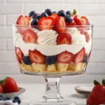 Close-up of a 4th of July trifle in a clear glass trifle bowl, layered with angel food cake cubes, whipped topping, strawberries, and blueberries, surrounded by fresh berries.