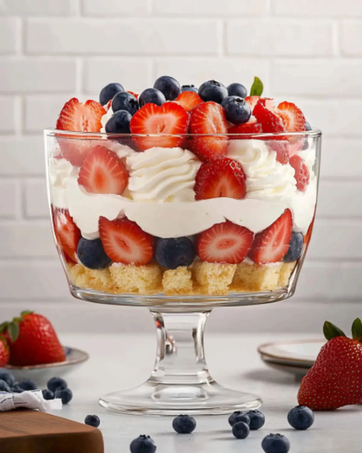 Close-up of a 4th of July trifle in a clear glass trifle bowl, layered with angel food cake cubes, whipped topping, strawberries, and blueberries, surrounded by fresh berries.