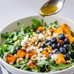 Honey lemon vinaigrette being drizzled over honey sweet potato summer salad in a white bowl with roasted sweet potatoes, arugula, blueberries, goat cheese, pepitas, and pine nuts.