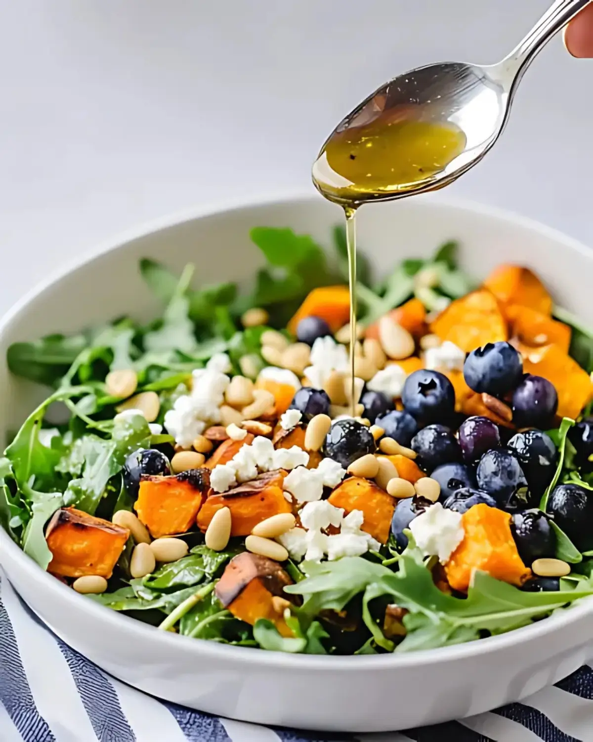 Honey lemon vinaigrette being drizzled over honey sweet potato summer salad in a white bowl with roasted sweet potatoes, arugula, blueberries, goat cheese, pepitas, and pine nuts.