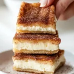 Close-up of flaky Churro Cheesecake Bars stacked on a white plate with cinnamon sticks, perfect for an easy cinnamon dessert.