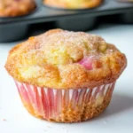 Close-up of a freshly baked rhubarb muffin topped with cinnamon sugar, sitting in front of a muffin tin on a white surface
