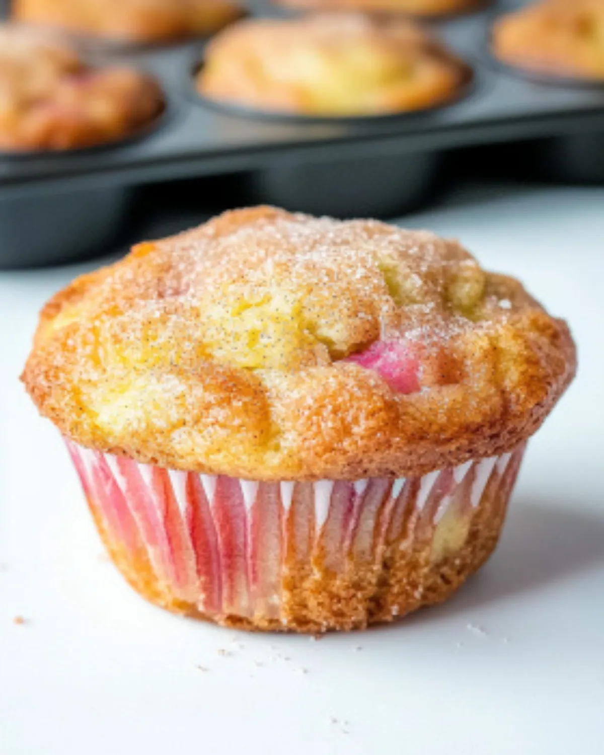 Close-up of a freshly baked rhubarb muffin topped with cinnamon sugar, sitting in front of a muffin tin on a white surface
