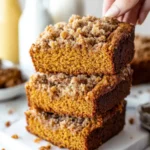 Stack of three slices of Moist Pumpkin Bread with streusel topping, one being lifted by hand, with blurred milk bottles in the background.