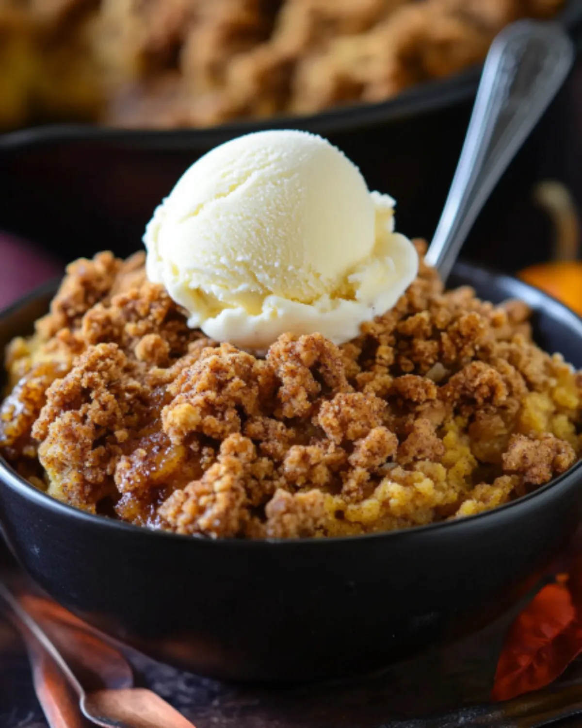 Single serving of pumpkin crisp in a black bowl with melting vanilla ice cream on top and a spoon ready to dive in.