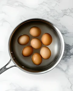 Brown eggs in stainless steel pot filled with water on marble countertop before boiling