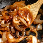 Wooden spoon scooping rich, golden caramelized onions from a cast iron skillet, glistening in oil.