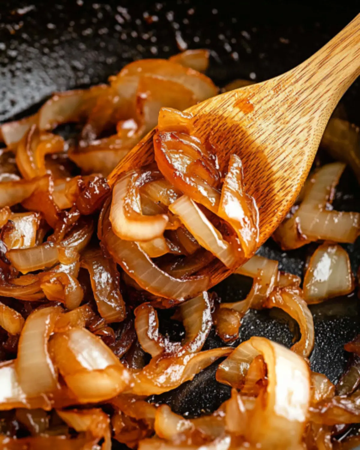 Wooden spoon scooping rich, golden caramelized onions from a cast iron skillet, glistening in oil.