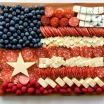 American Flag Charcuterie Board with blueberries, raspberries, salami, white cheese cubes, crackers, and a large star-shaped cheese centerpiece on a wooden board