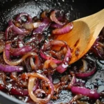 Rich and glossy Red Wine Onions cooking down in a black skillet, stirred with a wooden spoon.