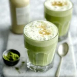 Two glasses of frothy iced matcha latte with almond milk on marble tray, surrounded by matcha powder and milk bottle