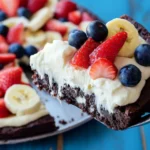 Brownie dessert pizza with cream cheese frosting, strawberries, bananas, and blueberries on a rustic blue background.