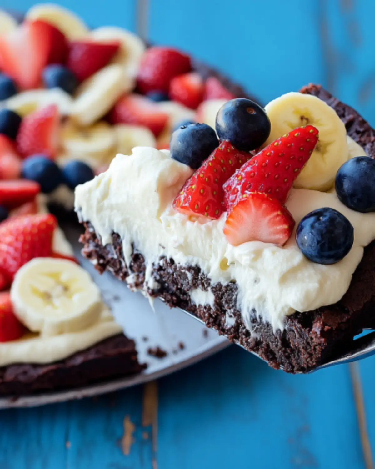 Brownie dessert pizza with cream cheese frosting, strawberries, bananas, and blueberries on a rustic blue background.