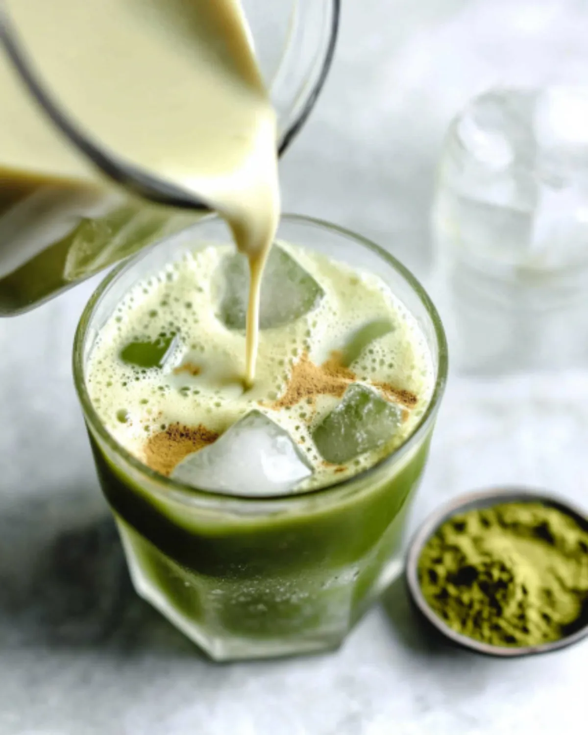 Glass of iced matcha latte with almond milk being poured, surrounded by matcha powder on a marble surface