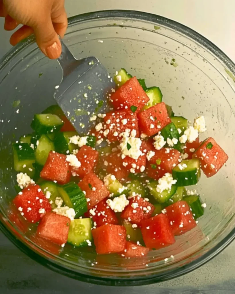 A hand gently tossing a Watermelon Salad With Cucumber And Feta using a spatula. The fresh ingredients include watermelon cubes, cucumber slices, and crumbled feta.