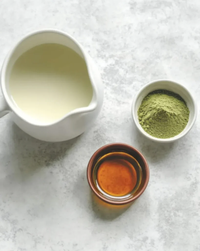 Iced matcha latte ingredients including almond milk, ceremonial grade matcha powder, and 0 calorie vanilla syrup on a light marble surface