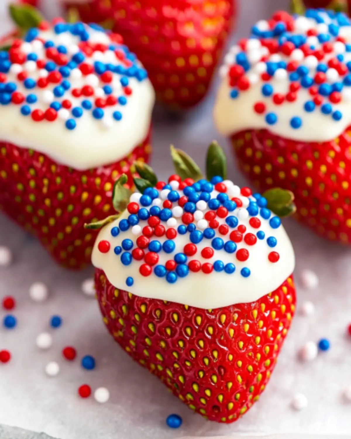 Close-up of a chocolate-covered strawberry with white chocolate and red, white, and blue sprinkles on white paper.