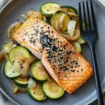 Top-down view of baked sesame-ginger salmon fillet topped with black and white sesame seeds, served over sautéed zucchini and onions on a gray plate