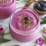 Vibrant berry smoothie topped with chia and flax seeds in a round glass, surrounded by blueberries and flowers