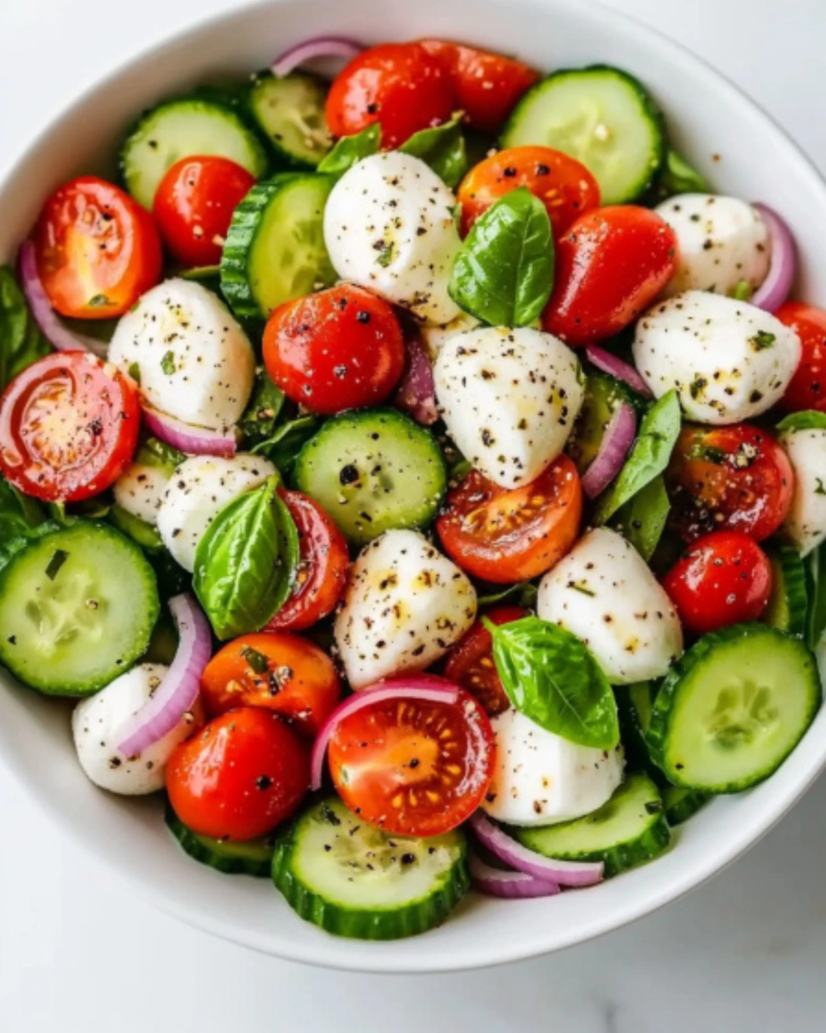 Close-up of cucumber Caprese salad with red onion, mozzarella, tomatoes, and chopped herbs in a white bowl.
