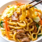 Chopsticks lifting stir-fried Yaki Udon noodles with pork and vegetables from a white bowl