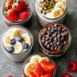 Five jars of chia pudding topped with chocolate chips, banana slices, and fresh strawberries on a gray background