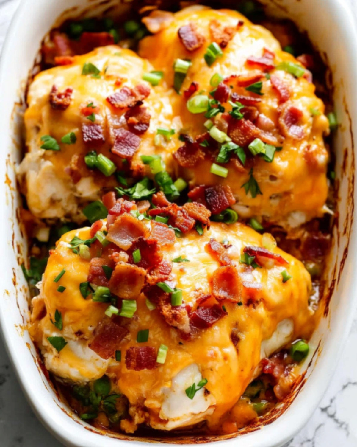 Baked crack chicken topped with crispy bacon, melted cheddar cheese, and green onions in a white casserole dish