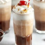 Tall glass of chocolate Italian cream soda with whipped cream, cherry, and red-striped straw on a white surface.