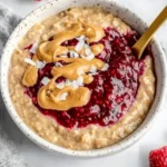 Creamy peanut butter and jelly oatmeal in a white bowl with chia jam and coconut flakes, gold spoon inside.