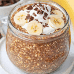 Creamy chocolate banana oats in a glass mug, topped with whipped cream, chocolate, and banana slices.