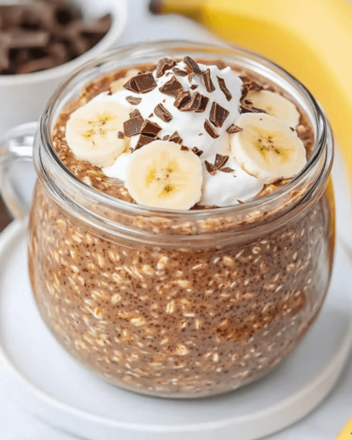 Creamy chocolate banana oats in a glass mug, topped with whipped cream, chocolate, and banana slices.