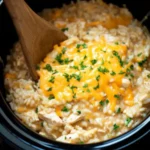 Creamy crockpot chicken and rice topped with melted cheddar and parsley in a black slow cooker with wooden spoon