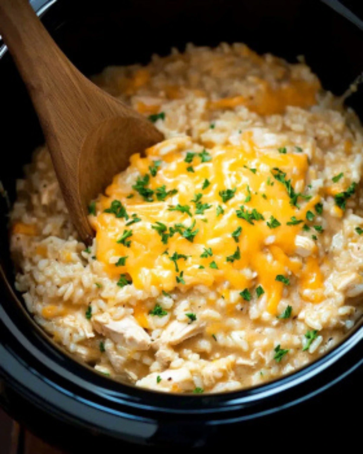 Creamy crockpot chicken and rice topped with melted cheddar and parsley in a black slow cooker with wooden spoon
