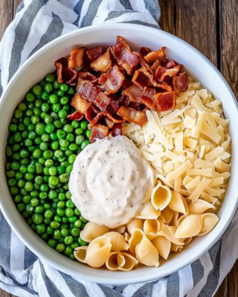 Overhead view of creamy pasta salad ingredients in a bowl: shell pasta, peas, bacon, parmesan, and mayo dressing