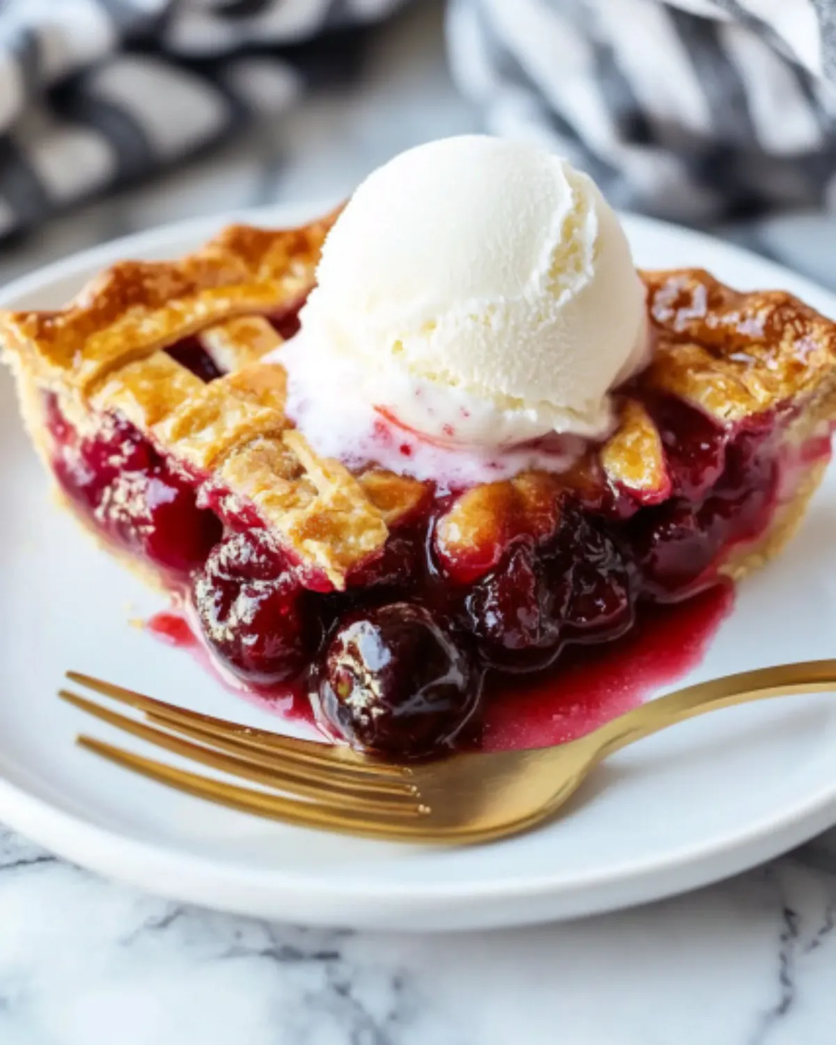 Slice of homemade cherry pie with golden crust and vanilla ice cream on a white plate with a gold fork