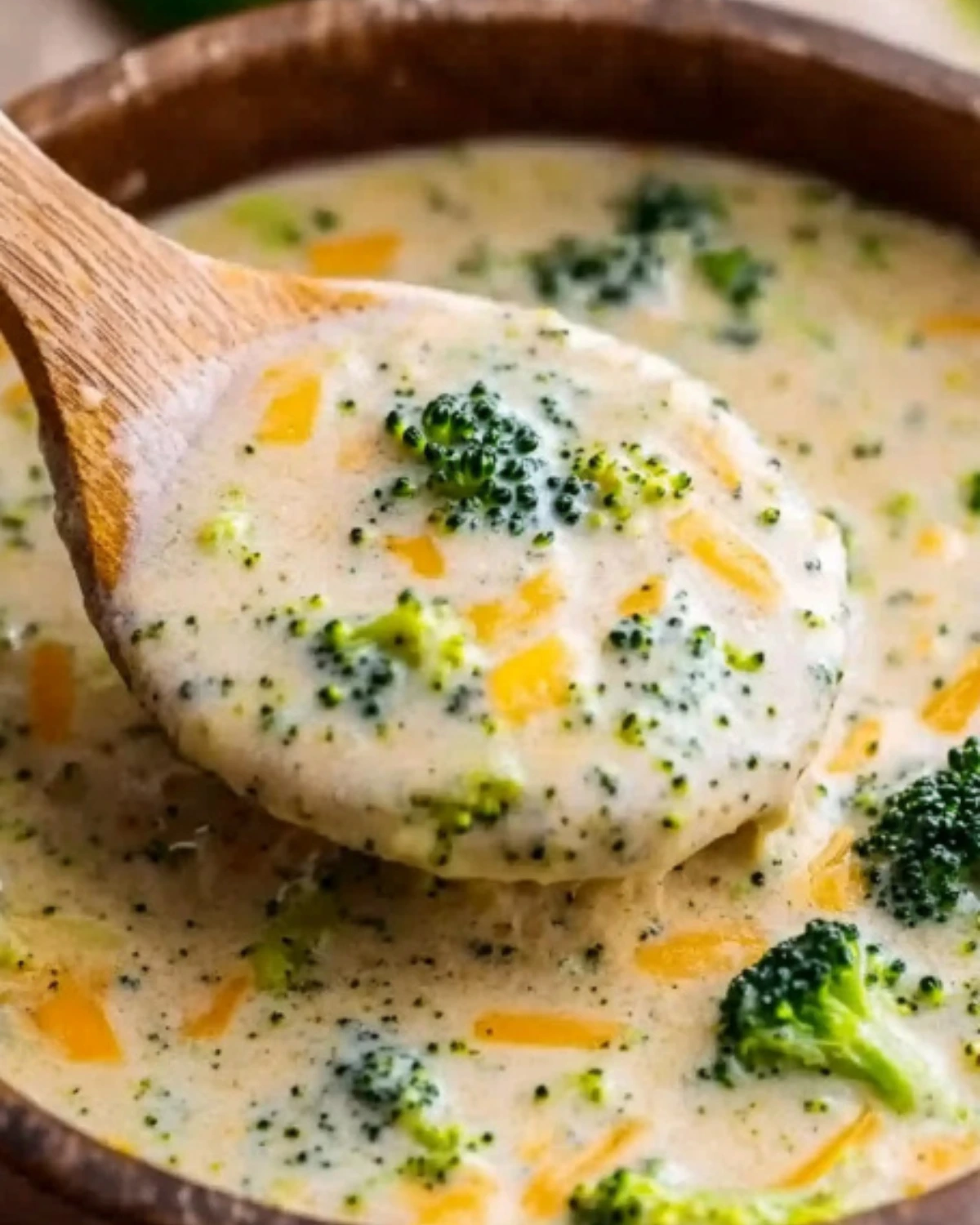 A wooden spoon scooping creamy broccoli cheddar soup with visible broccoli florets and melted cheddar cheese, served in a rustic bowl.