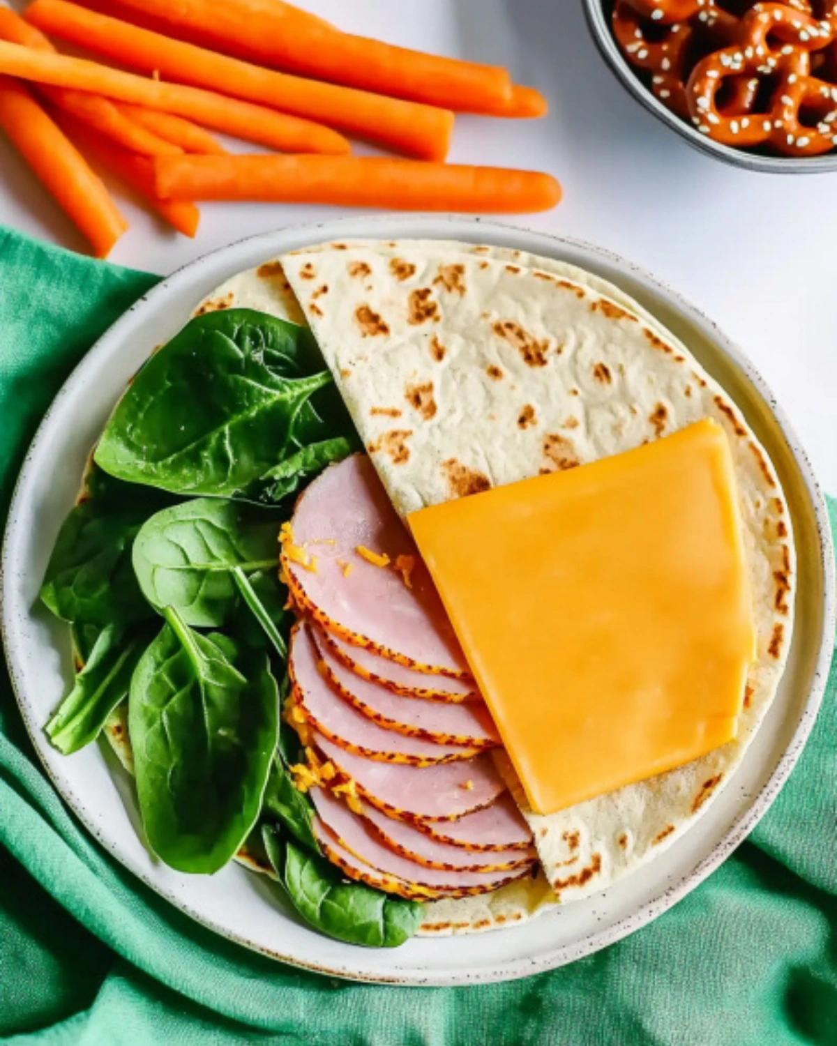 Tortilla wrap with spinach, slices of ham, cheddar cheese, and a side of carrot sticks and pretzels.