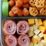A healthy and visually appealing lunchbox with deli ham slices, cheese cubes, grapes, pretzel crisps, and snap peas in a clear glass container.