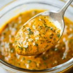 Close-up of vibrant yellow-orange marinade with herbs on a spoon in a clear glass bowl.