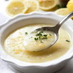 A close-up of creamy lemon garlic butter sauce in a white dish with a spoon, garnished with fresh parsley and surrounded by lemon slices.