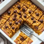 Pumpkin baked oatmeal sliced in a white baking dish with chocolate chips and a fork lifting one square