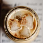 Overhead view of iced coffee with large ice cubes and a sprinkle of cinnamon, served in a mason jar with handwritten notes in the blurred background.